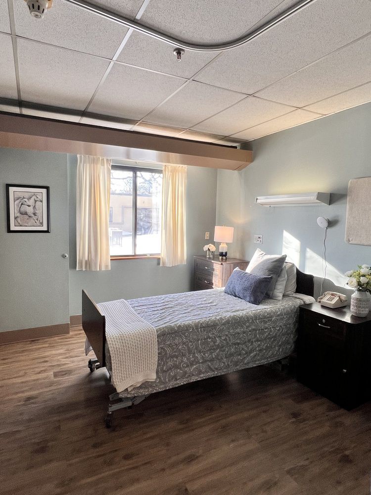 A hospital room with a single bed, neatly arranged with pillows and a blanket, next to a window with curtains. A nightstand holding a lamp and flowers is placed beside the bed.