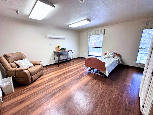 A spacious room featuring a bed, a comfortable chair, and wooden flooring with natural light coming through the window.