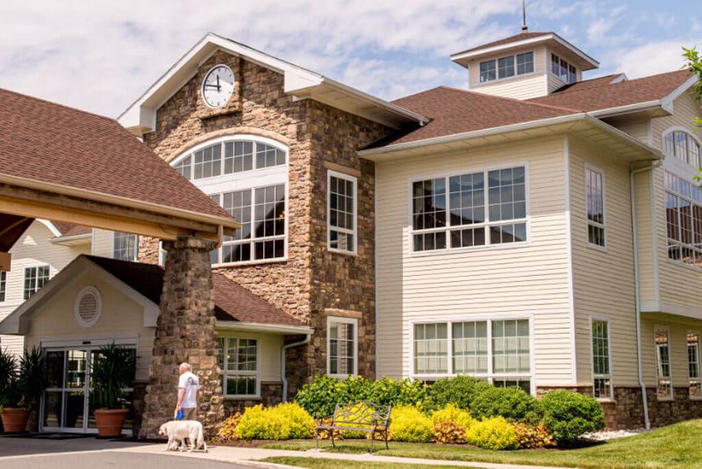 A person walking a dog outside a large residential building with stone and siding exterior.