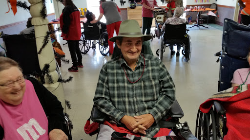 An elderly man in a plaid shirt and hat smiles while seated in a wheelchair during a social gathering.