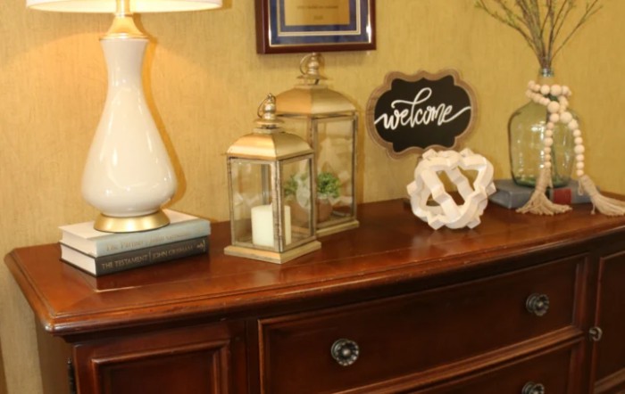 A warmly decorated table featuring a lamp, lanterns with candles, a 'welcome' sign, and a decorative ornament.