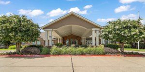 A modern building entrance with landscaped surroundings and a driveway under a clear sky.