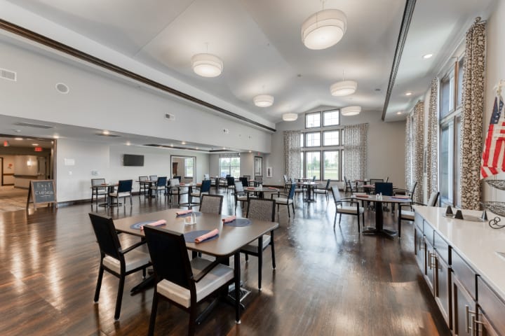 A spacious dining room with wooden floors, well-arranged tables and chairs, featuring large windows and modern lighting.