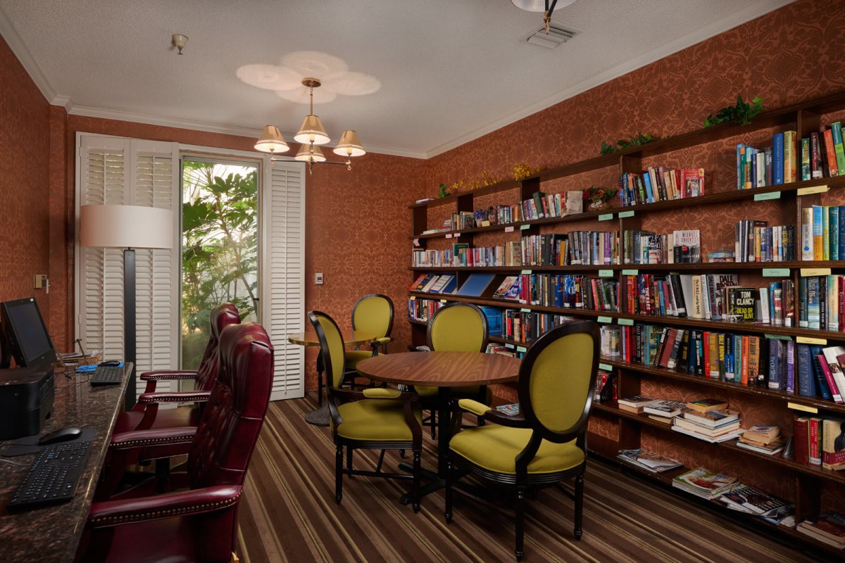 A cozy library with wooden floors, featuring bookshelves filled with books and comfortable seating around a table.