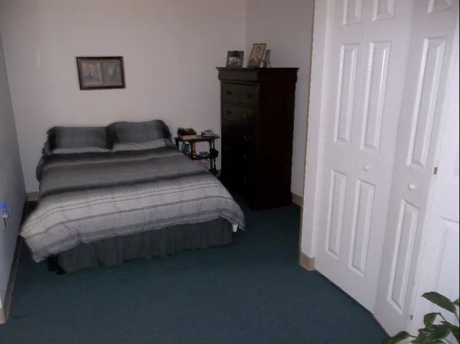 A simple bedroom with a neatly made bed, dresser, and carpeted floor.