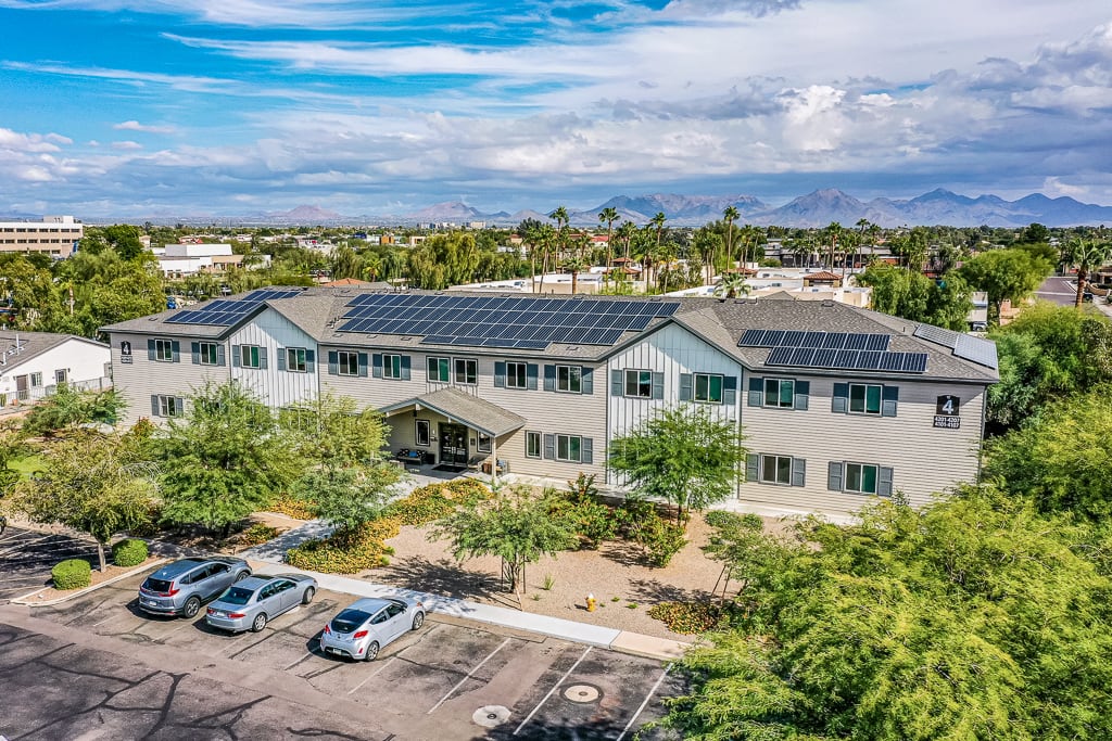 Aerial view of a building with solar panels on the roof, surrounded by trees and a parking lot under a cloudy sky.