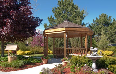 A wooden gazebo surrounded by a landscaped garden with trees and plants, set on a sunny day.