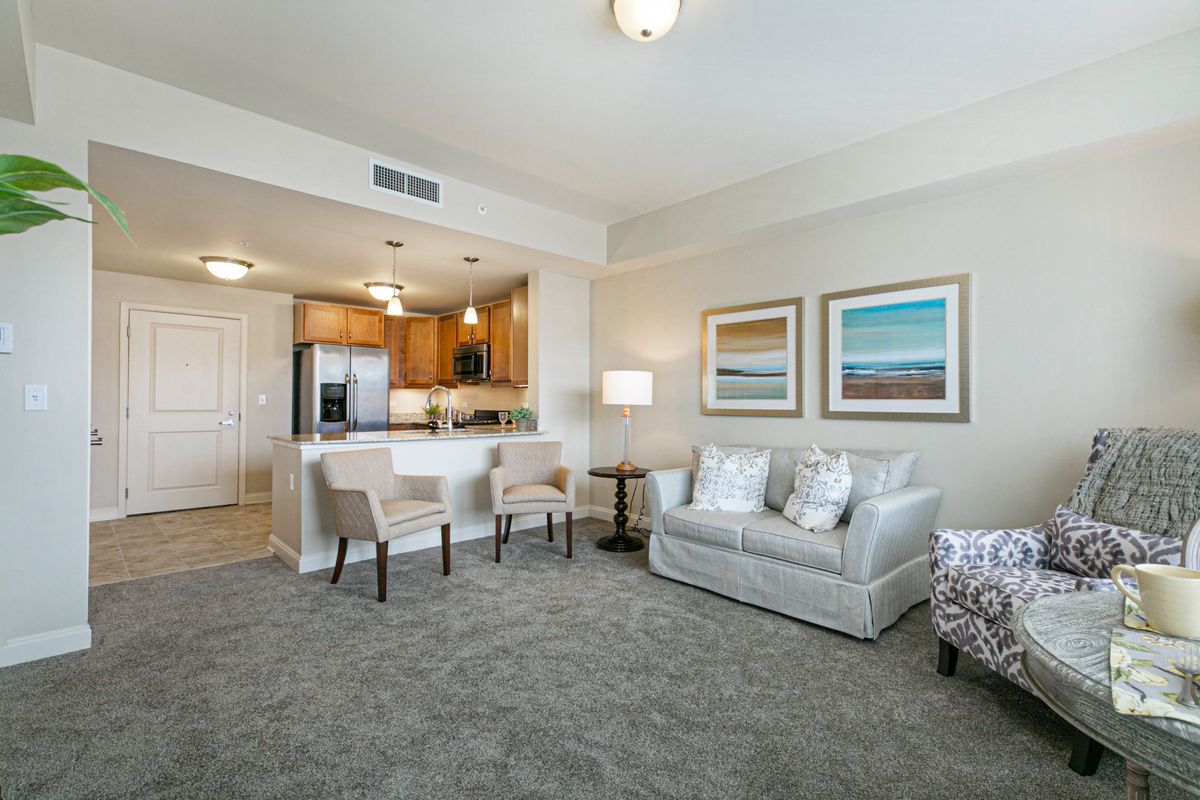 A modern living room with an open kitchen, featuring a sofa, bar stools, and artwork on the walls.