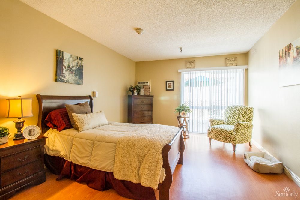 A cozy bedroom with a neatly made bed, wooden furniture, and ample natural light streaming through large windows.