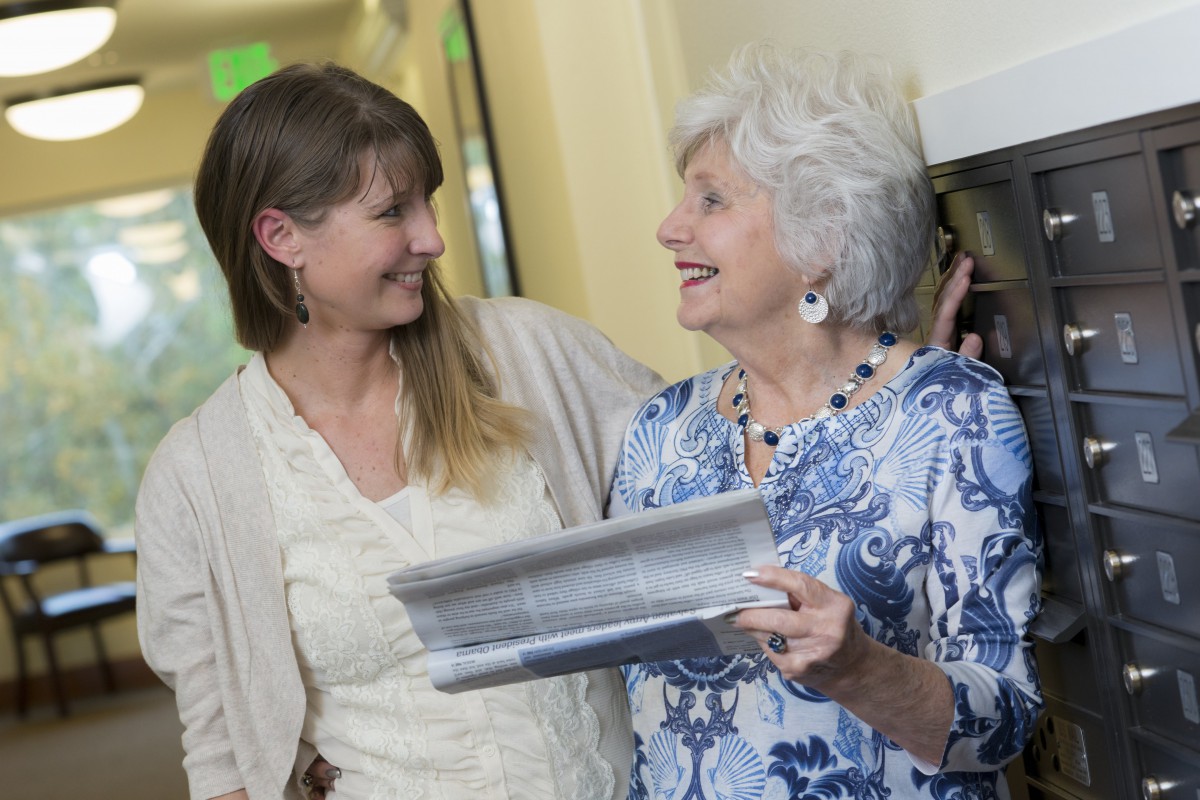 An elderly woman and a younger woman smiling together while holding a newspaper indoors.