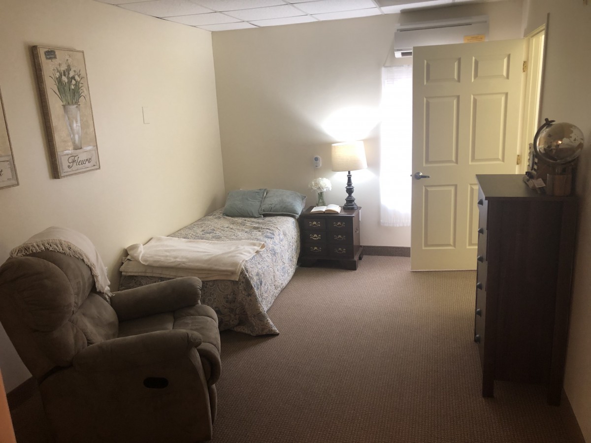 A simple bedroom with a single bed, armchair, dresser, and bedside lamp.