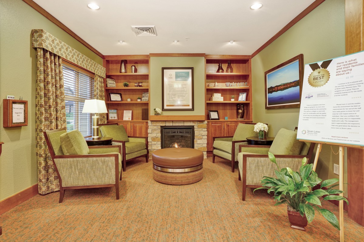 A cozy living room with comfortable seating around a fireplace, surrounded by bookshelves and decorated with plants and artwork.