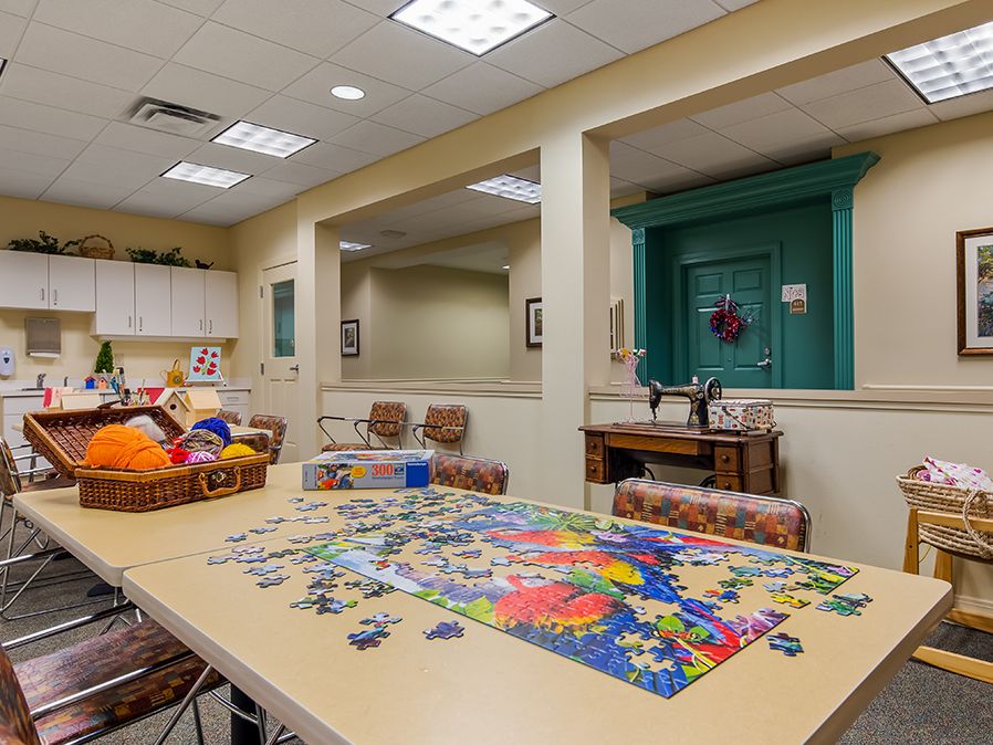 A brightly lit activity room featuring a large table with a partially completed puzzle, surrounded by chairs.