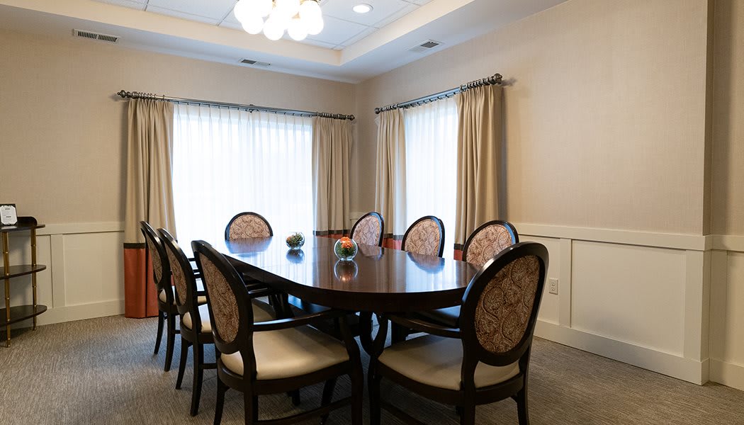 A formal dining room with an oval table surrounded by upholstered chairs and bright curtains.