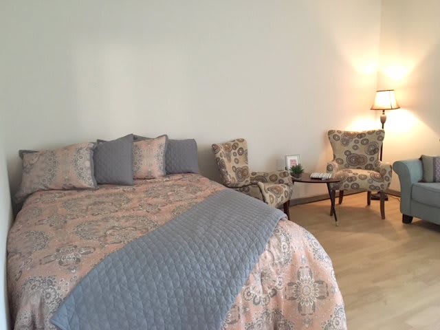 A cozy bedroom with a neatly made bed, floral chairs, and warm lighting.