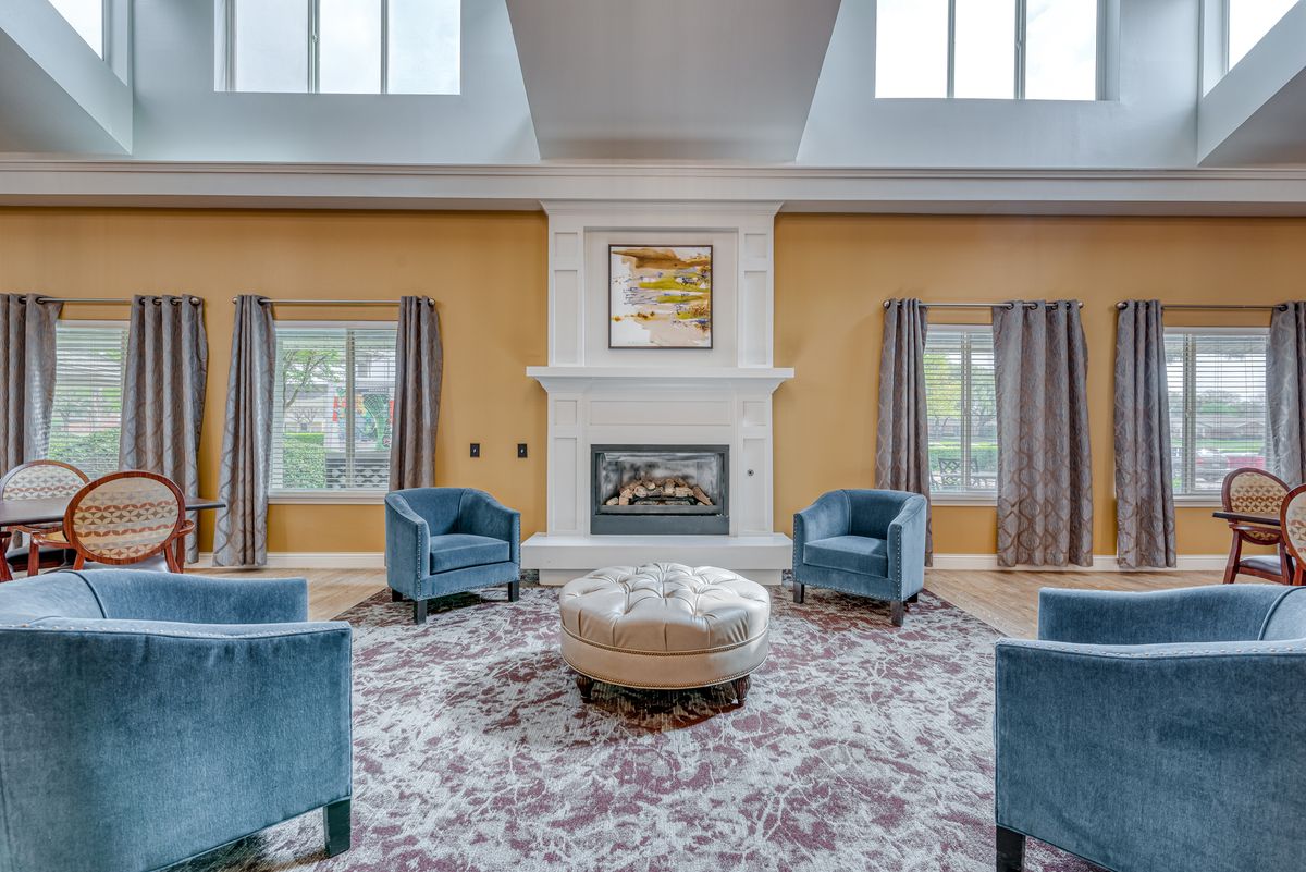 A spacious living room featuring a central fireplace, surrounded by blue chairs and a patterned carpet under natural light from large windows.