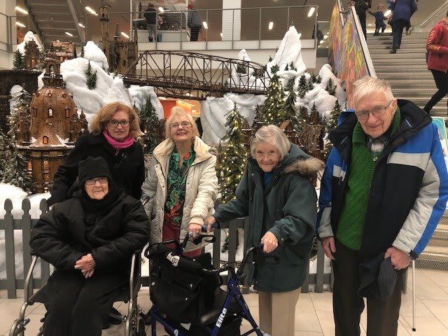 A group of elderly people gathered indoors, dressed in winter clothing and surrounded by holiday decorations.