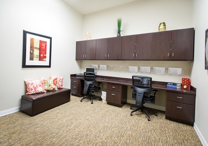 A tidy office workspace with two desks, chairs, and computers, featuring overhead cabinets and decorative items.