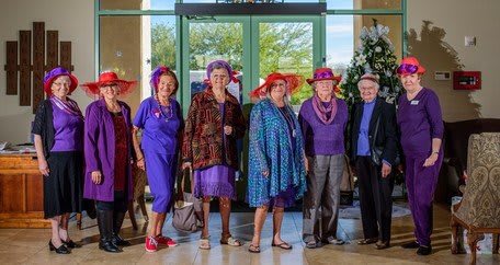 A group of people dressed in colorful outfits and hats gathers for a photo indoors.