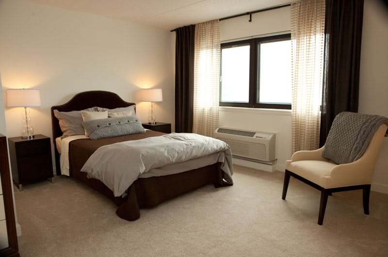 A cozy and modern bedroom with a neatly made bed, a bedside lamp, and an armchair near the window.