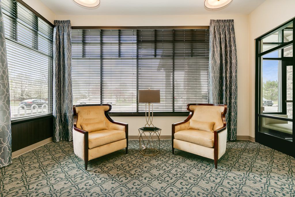 A cozy living room setup featuring two armchairs, a small table with a lamp, and large windows with elegant curtains.