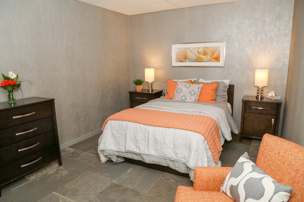 A cozy and modern bedroom with a neatly made bed featuring orange accent pillows, a lamp on the nightstand, and decorative wall art.
