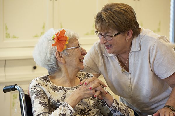 An elderly woman with a flower in her hair shares a warm moment with a caregiver.
