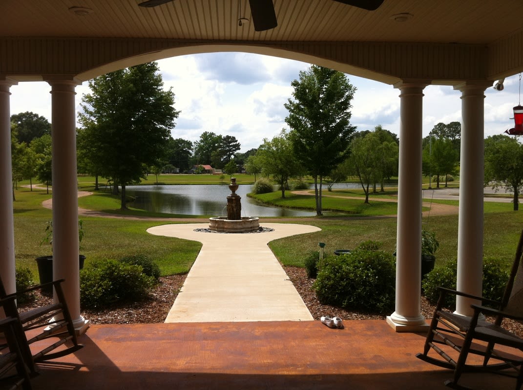 A serene view from a porch overlooking a garden with a statue, pathway, and pond surrounded by trees.
