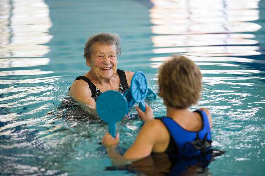 Two senior women engaging in aquatic therapy exercises in a swimming pool, using paddles to enhance their workout.