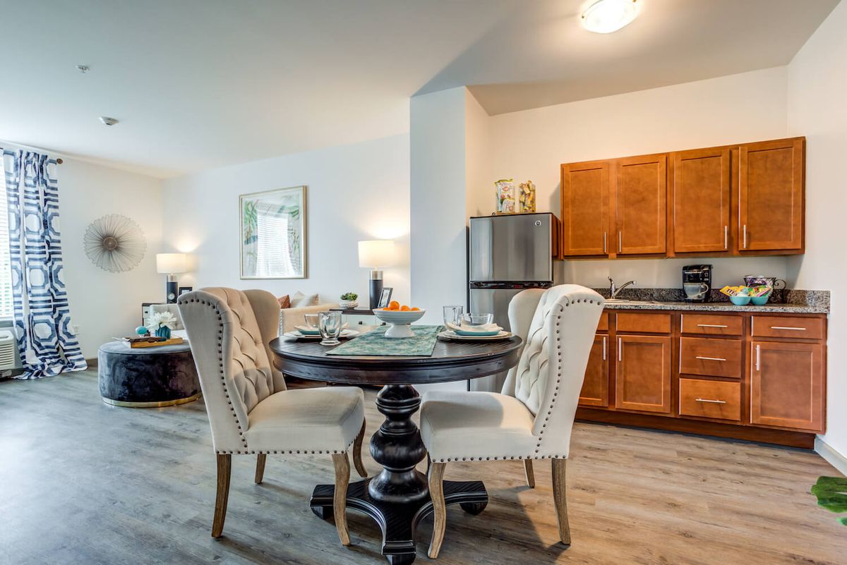 A modern dining area with a round table and upholstered chairs adjacent to a kitchen in an apartment setting.