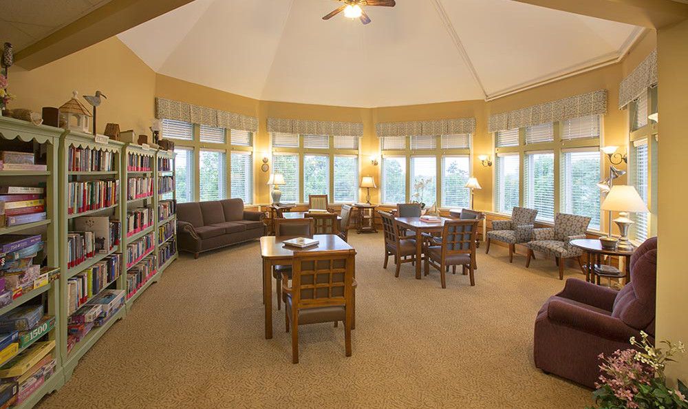 A cozy library reading room with bookshelves, tables, and chairs by large windows.