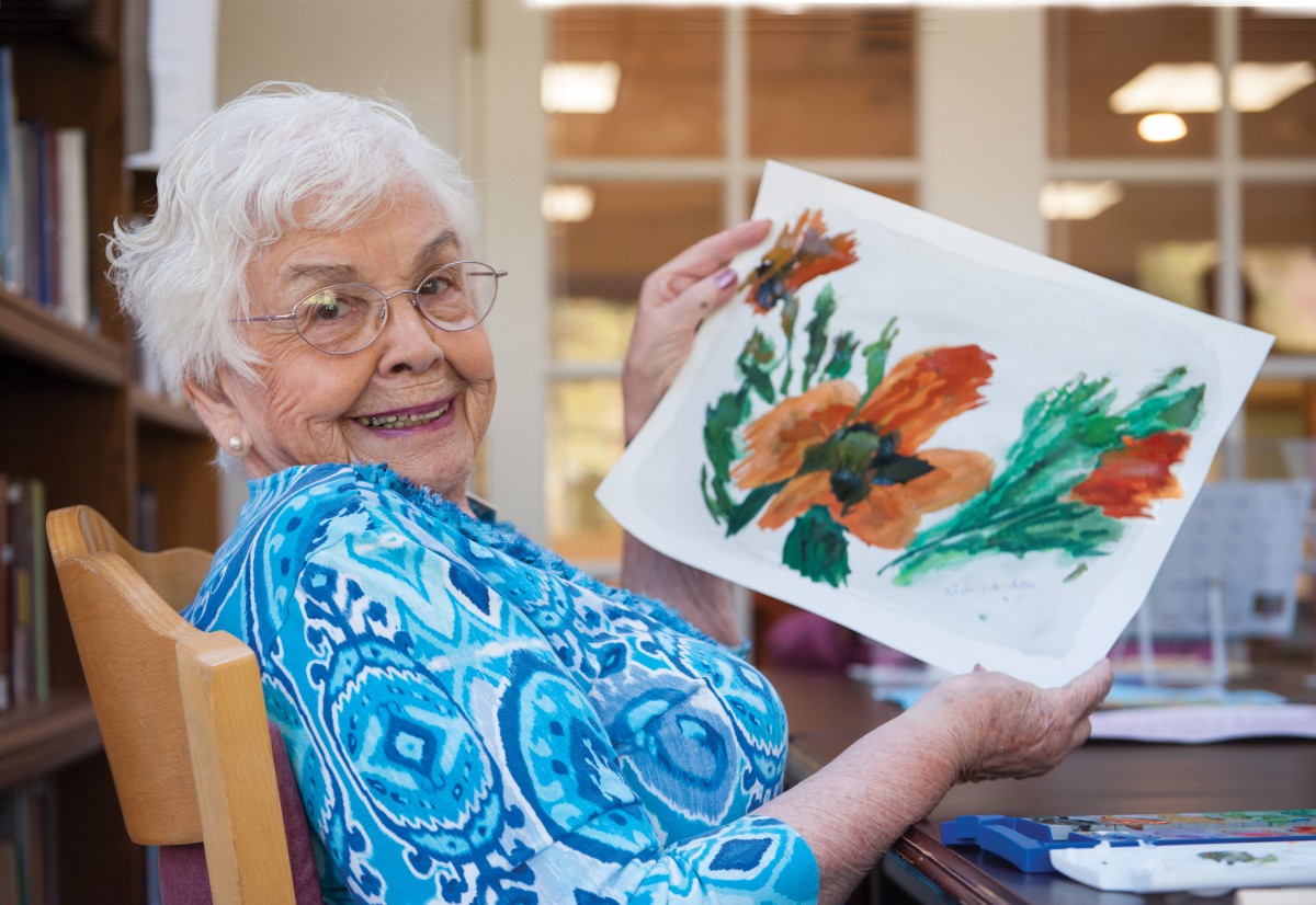 An elderly woman proudly displays her floral artwork.