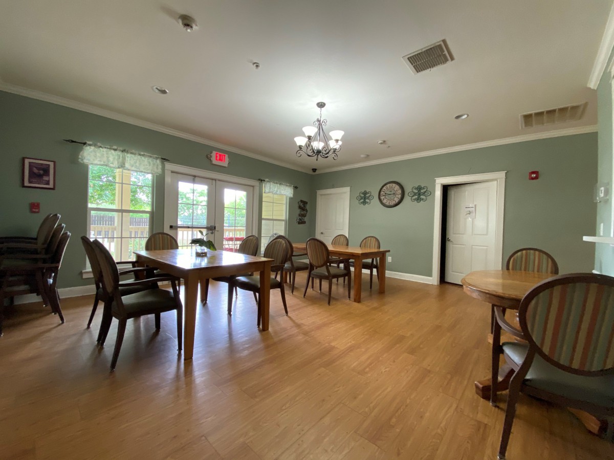 A spacious dining room with wooden floors and multiple tables and chairs, illuminated by a chandelier.