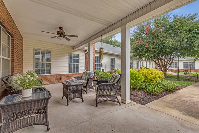 A cozy patio with wicker furniture and a ceiling fan, surrounded by a garden.