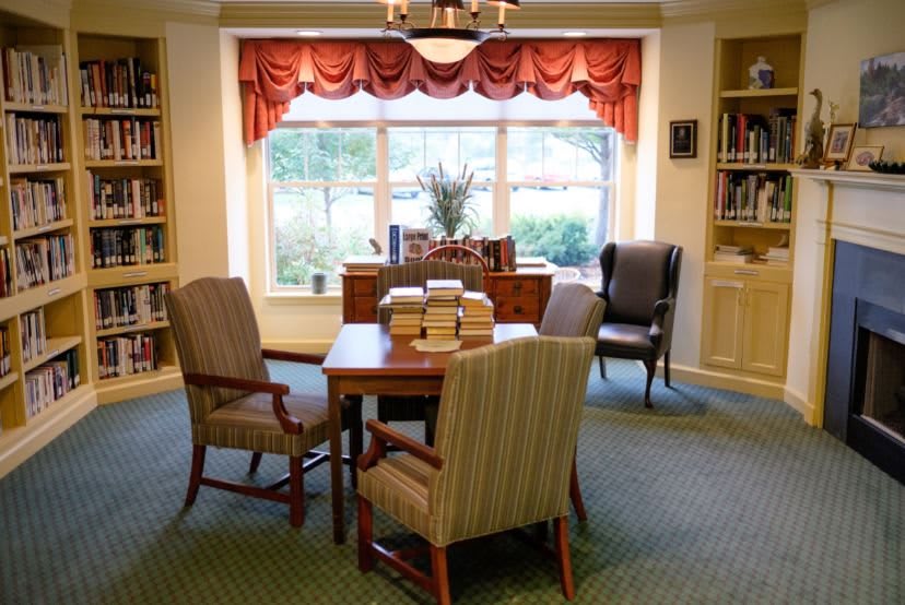 A cozy library reading area with a table, chairs, and stacks of books near a large window.