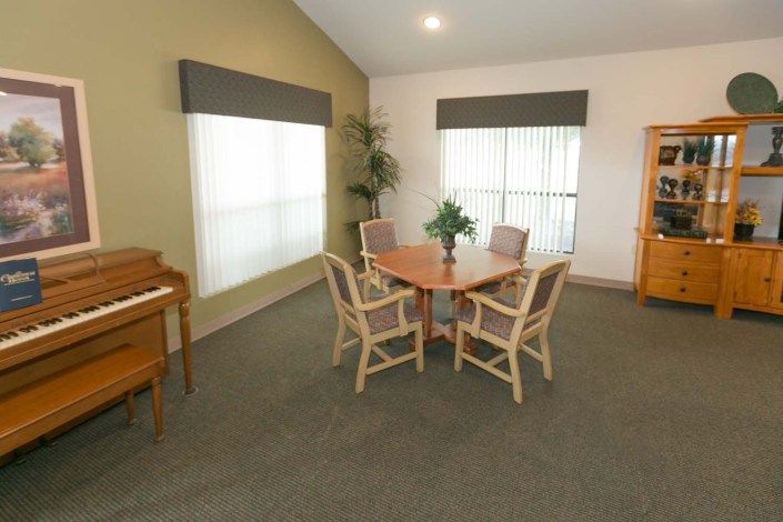 A cozy dining area with a round table and four chairs, complemented by large windows with blinds, an upright piano, and a potted plant in the corner.