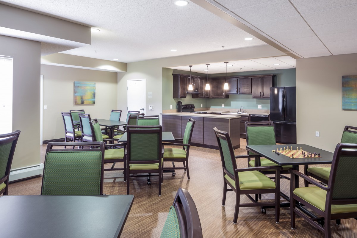 A well-lit dining area with green upholstered chairs and a modern kitchen in the background.
