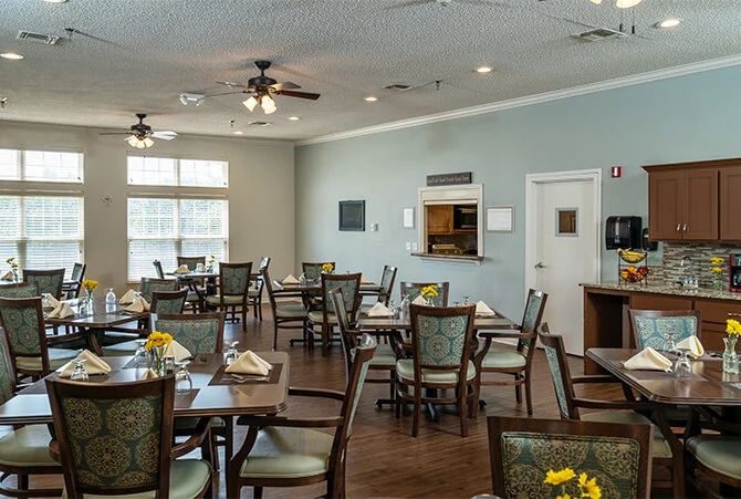 A spacious dining area with neatly arranged tables and chairs, featuring large windows and ceiling fans.