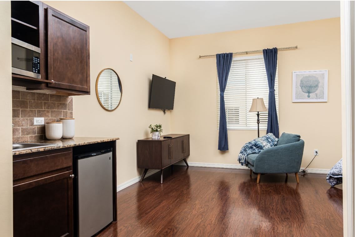 A cozy living room with wood flooring, featuring a sofa, television, and a small kitchen area with cabinets.