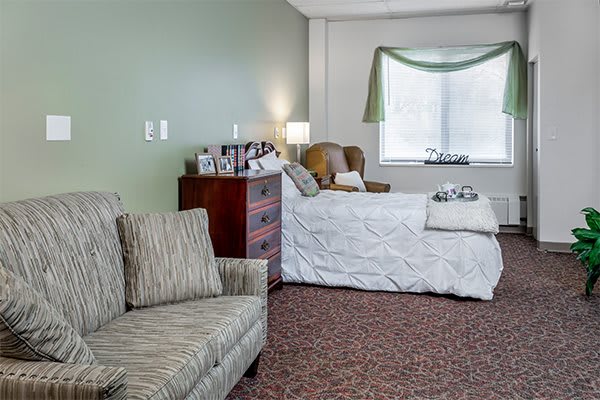 A cozy bedroom with a bed, dresser, and armchair near a window.