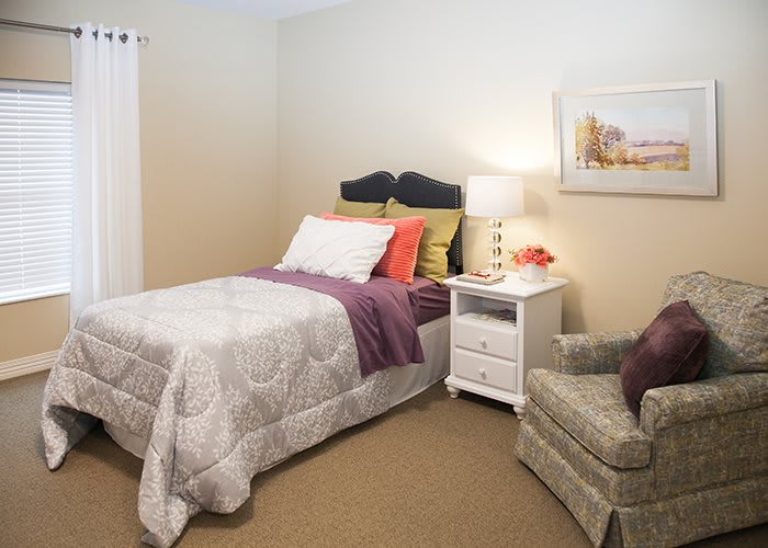 A cozy bedroom with a neatly made bed, a lamp on the nightstand, and an upholstered chair.