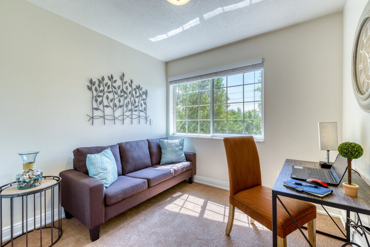 A cozy living room with a sofa, desk, and chair, illuminated by natural light from a large window. Wall art adds a decorative touch.
