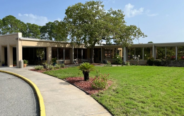 A single-story building surrounded by grass and trees, with a curved pathway leading to the entrance.