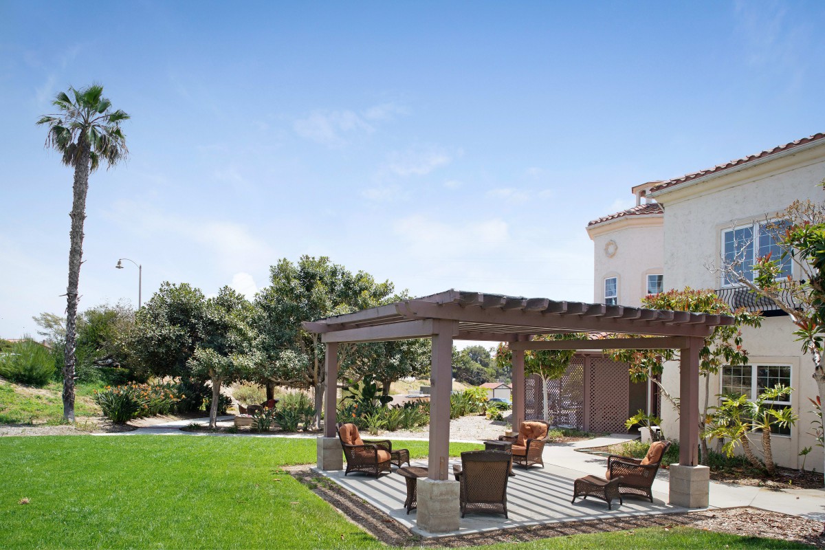 A serene backyard features a pergola with comfortable patio furniture, surrounded by a lush lawn and trees under a clear blue sky.