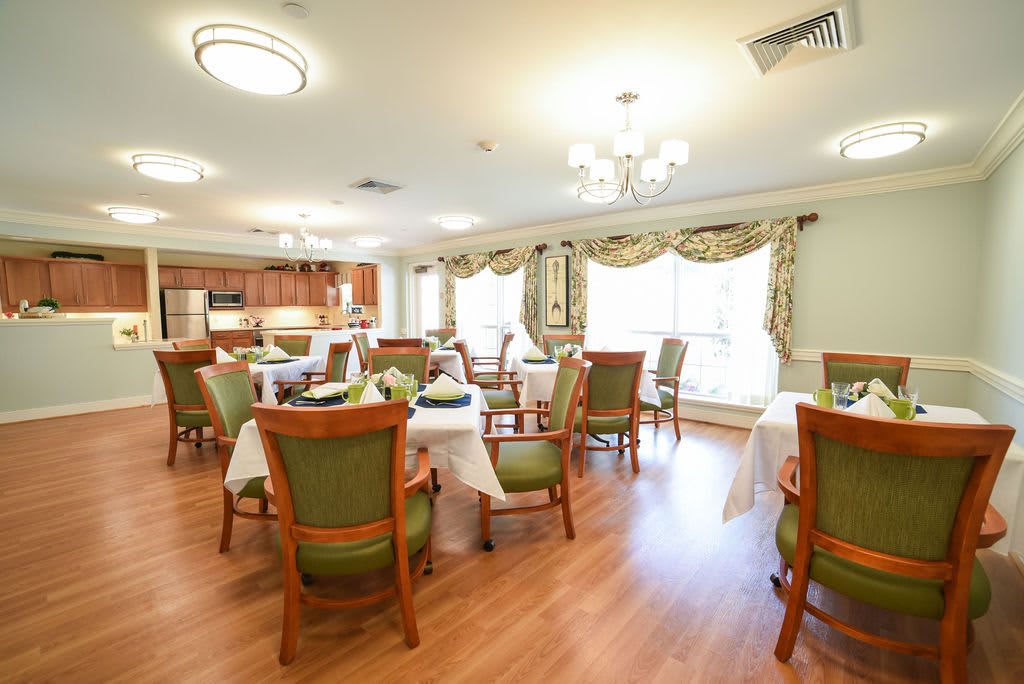 A well-lit dining room with elegantly set tables and chairs, featuring chandeliers and a kitchen area.