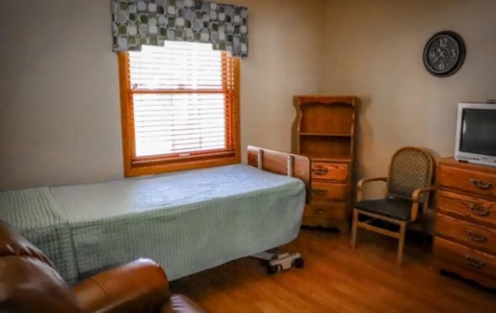 A small bedroom with minimal furnishings, featuring a bed, wooden shelves, and a window with blinds.