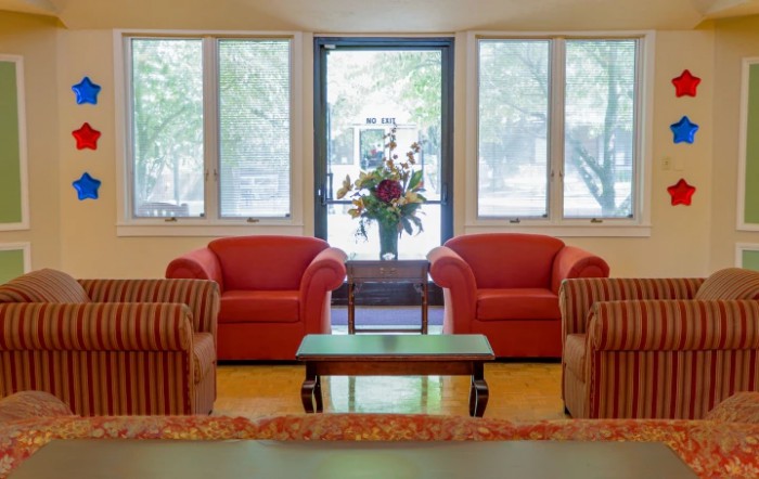 A cozy living room with two red armchairs, a central table, and large windows letting in natural light.