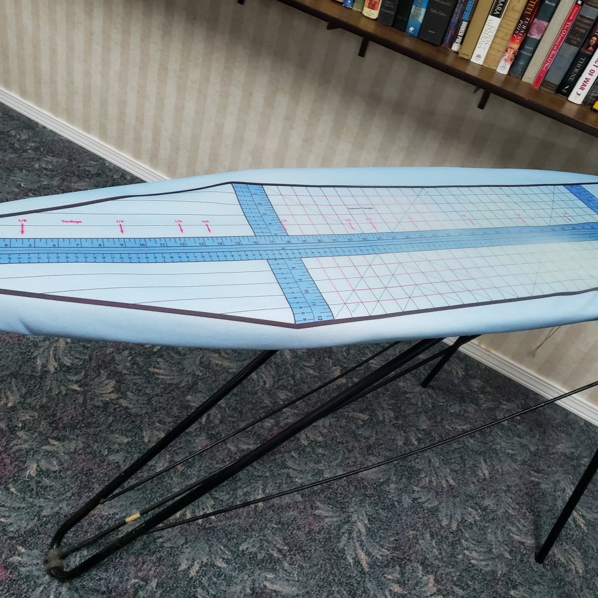 A blue ironing board set up in a room with carpeted flooring and bookshelves.