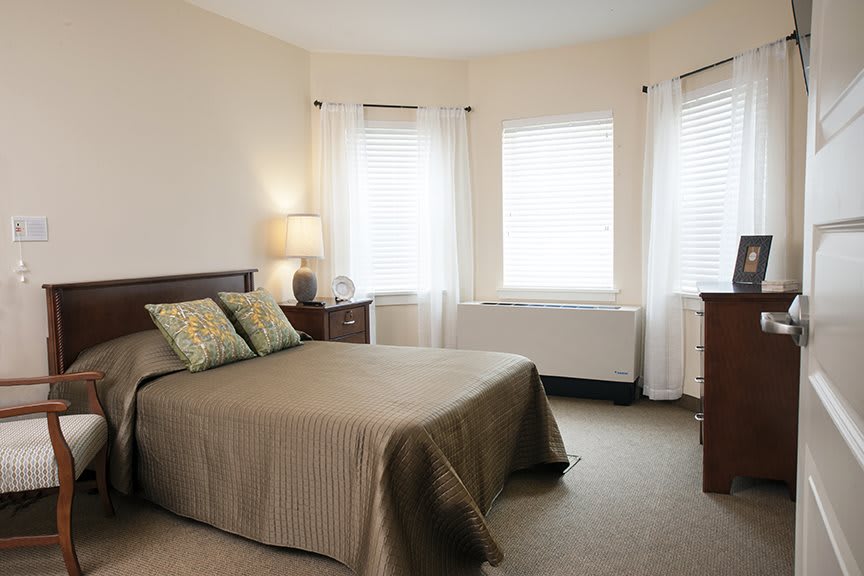 A cozy bedroom with a neatly made bed, bedside lamp, and ample natural light from the windows.