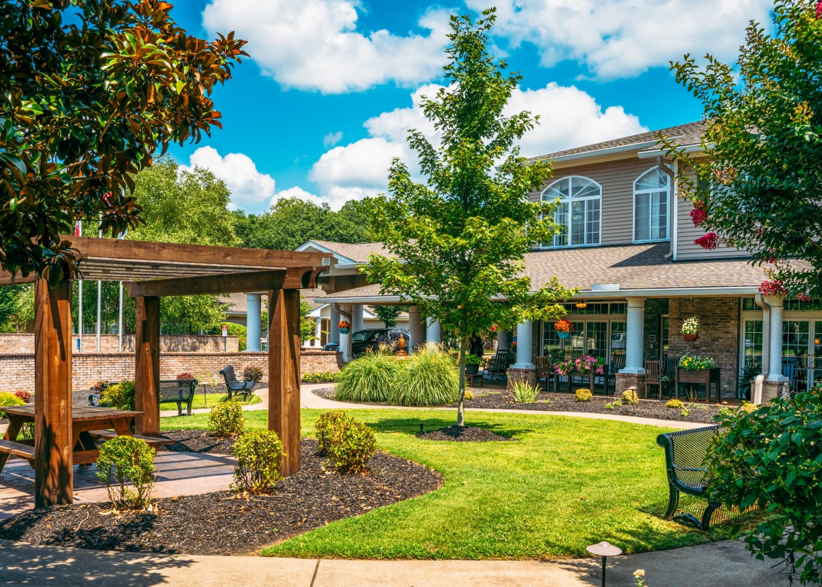 A beautifully landscaped garden with a wooden pergola and outdoor seating adjacent to a building under a bright blue sky.